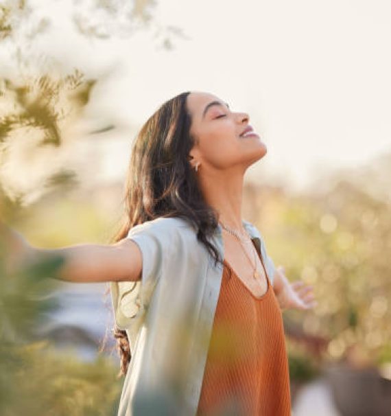 Young latin woman with arms outstretched breathing in fresh air during sunrise at the balcony. Healthy girl enjoying nature while meditating during morning with open arms and closed eyes. Mindful woman enjoying morning ritual while relaxing in outdoor park.