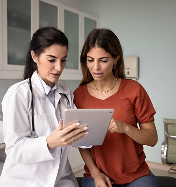 Focused young Latin doctor and female patient watching ultrasound scan shot on tablet, pointing at screen, talking, discussing examination result, medical report, electronic prescription